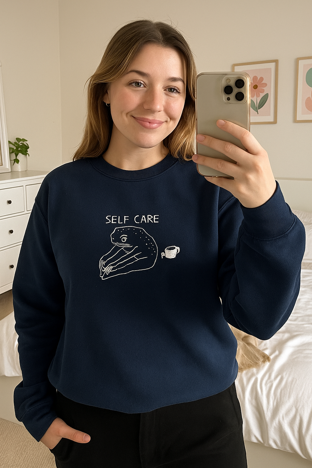 A woman is taking a selfie in a bedroom, wearing a navy blue sweatshirt with a white graphic and text that says "SELF CARE".