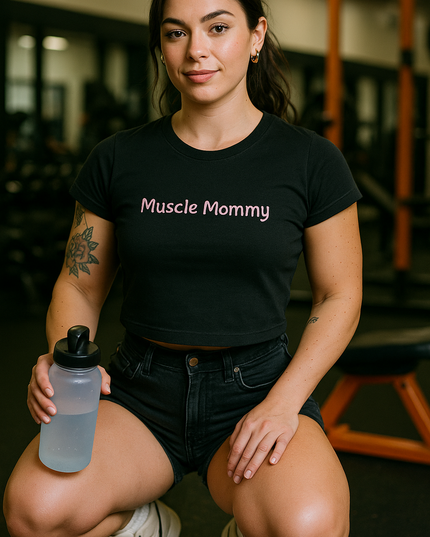 A woman wearing a black t-shirt with the text "Muscle Mommy" and black shorts is sitting on a bench in a gym, holding a water bottle.