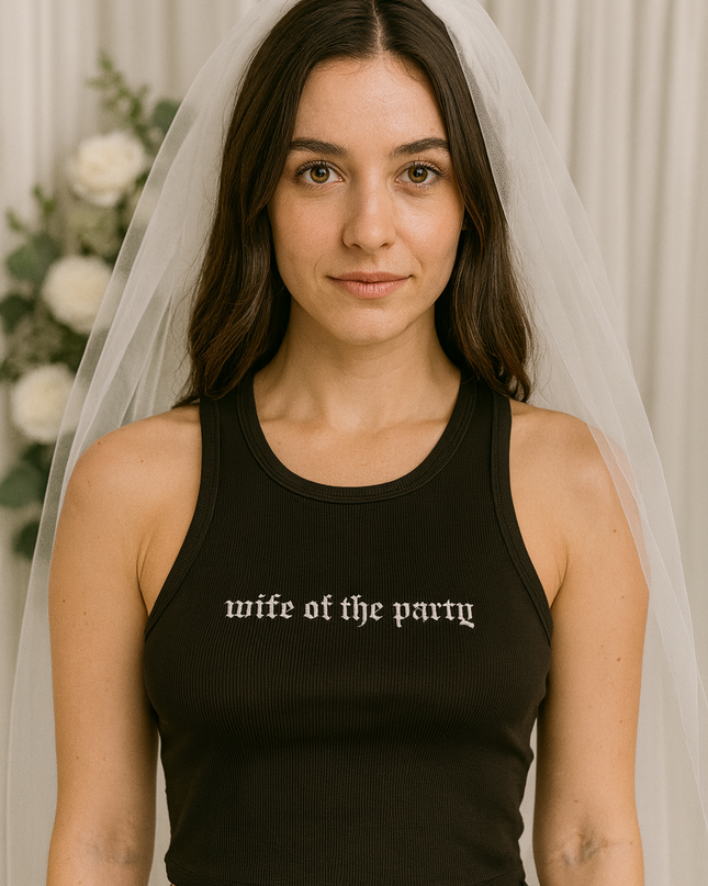 A woman wearing a black tank top with the text "wife of the party" printed on it, standing in front of a white curtain with white flowers.