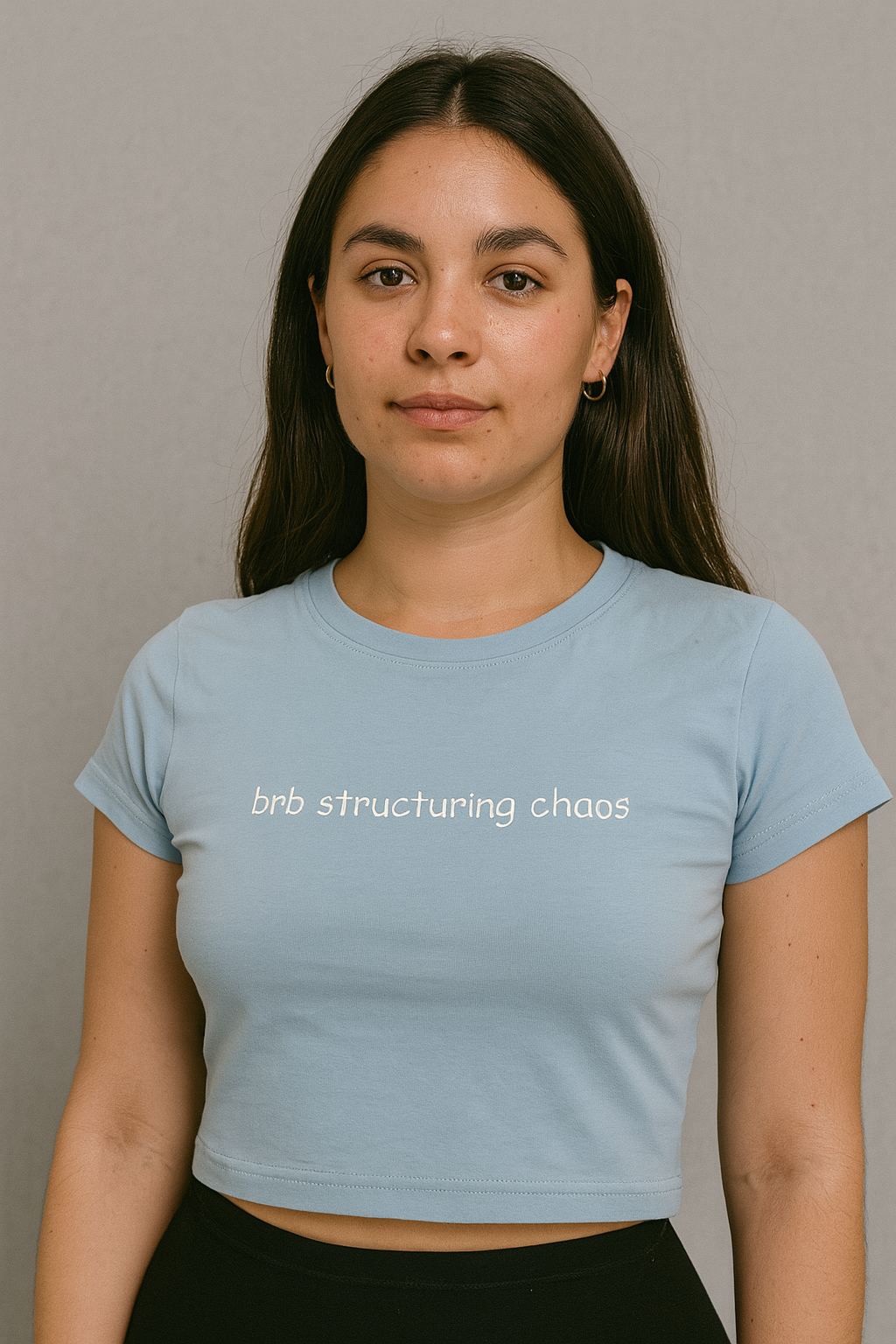 A young woman with long dark hair wearing a light blue crop top with the text "brb structuring chaos" printed on it.