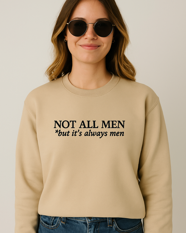 A woman wearing a beige sweater with the text "NOT ALL MEN *but it's always men" printed on it.