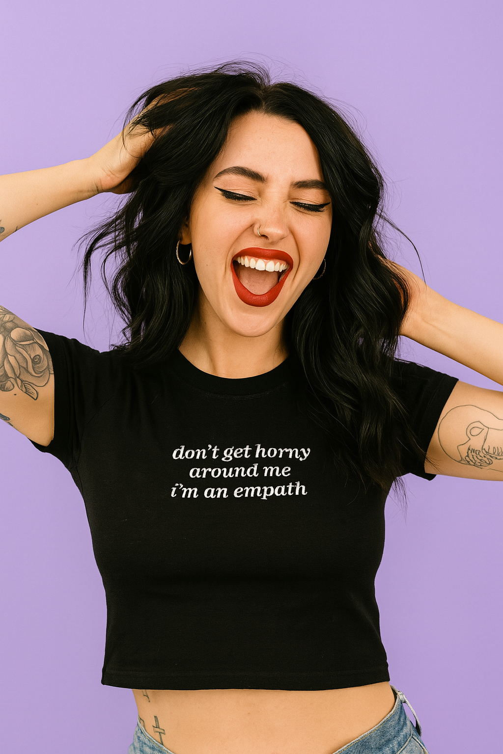 A woman with long dark hair is smiling and making a funny face, wearing a black crop top with white text.