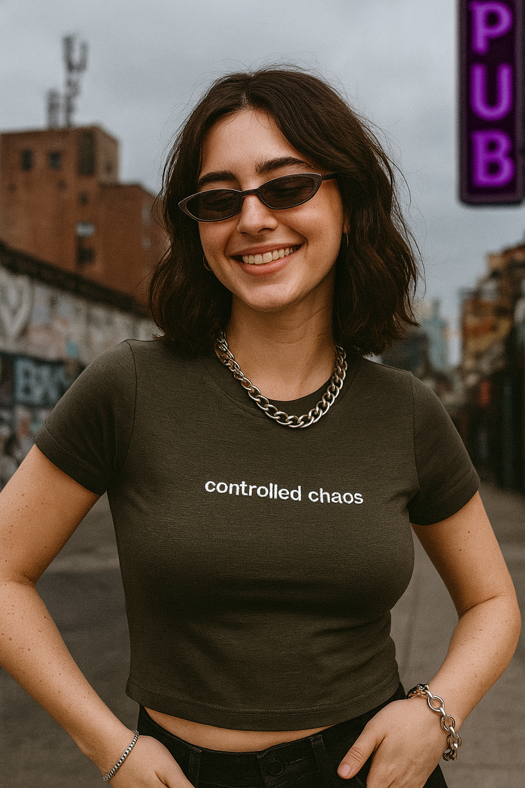 A smiling woman wearing a dark green t-shirt with the text "controlled chaos" printed on it, standing on a city street with graffiti-covered buildings in the background.