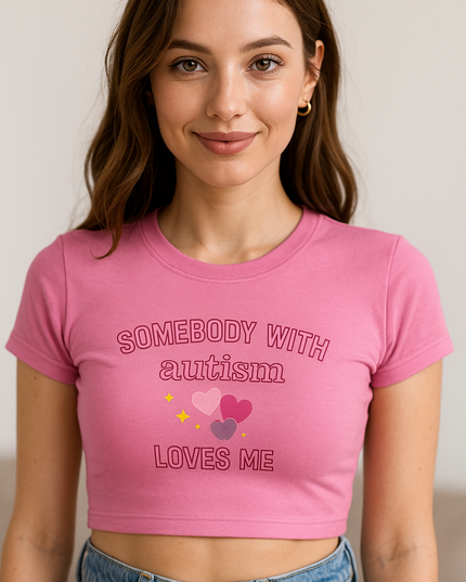 A young woman with long brown hair wearing a pink crop top with the text "SOMEBODY WITH AUTISM LOVES ME" printed on it.