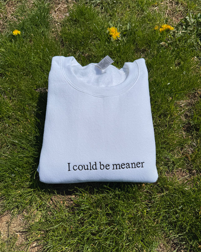A white t-shirt with the text "I could be meaner" printed on it, lying on a grassy surface with yellow flowers.
