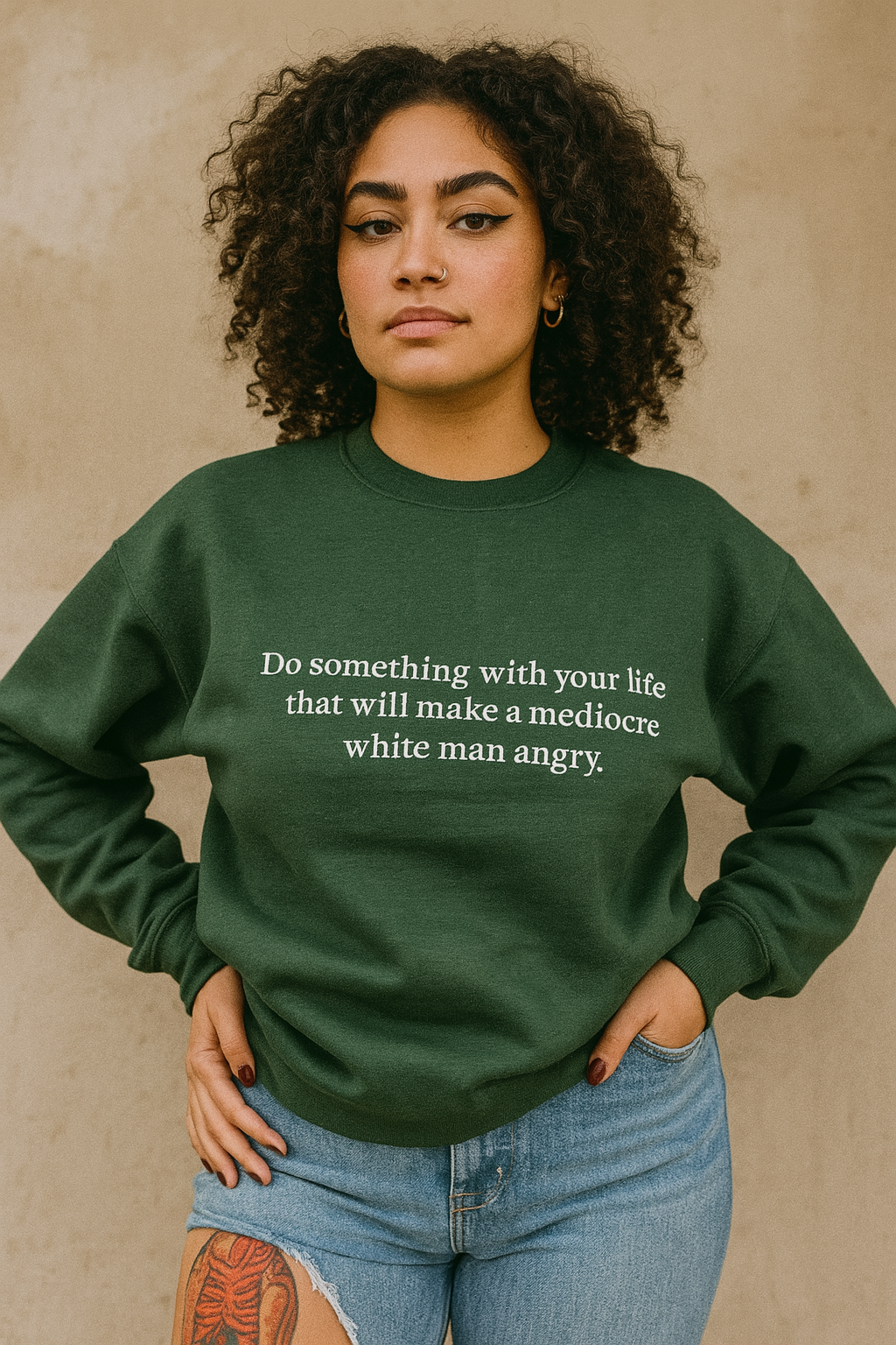 A young woman with curly hair wearing a green sweatshirt with a quote on it, standing against a beige wall.