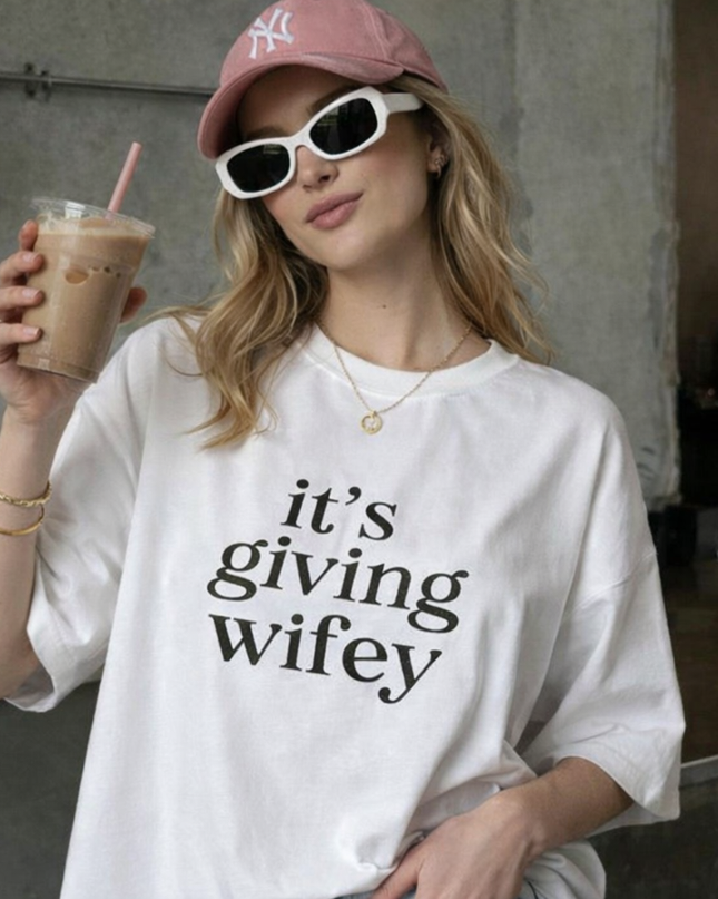 A woman wearing a white t-shirt with the text "it's giving wifey" and a pink cap is holding a drink with a straw.