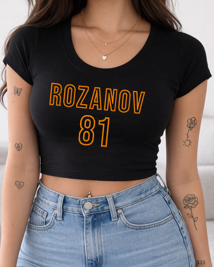 A woman wearing a black crop top with the text "ROZANOV 81" printed on it, paired with blue jeans. She has several tattoos on her arms and neck.