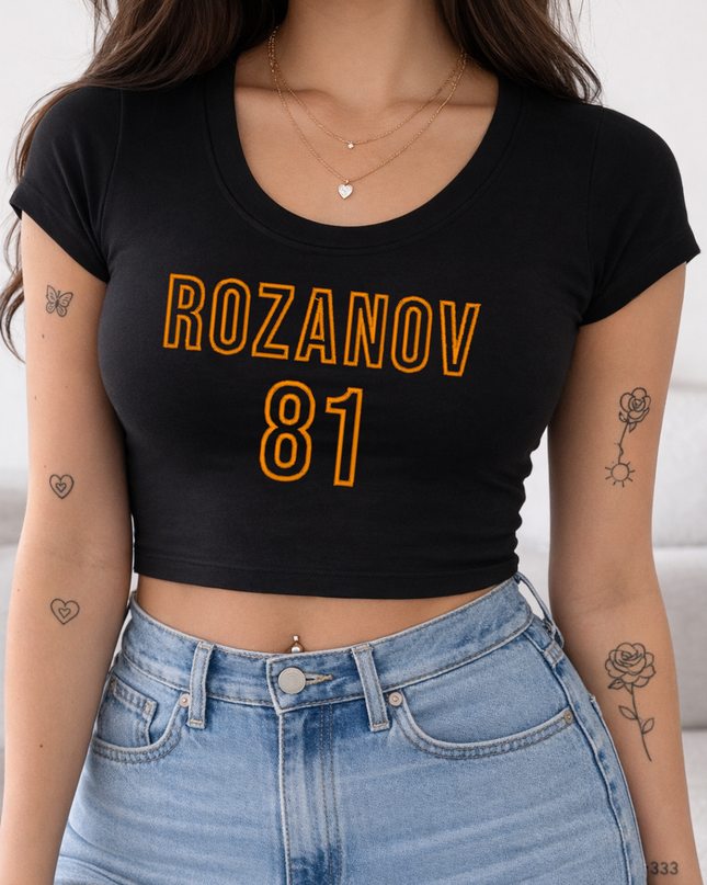 A woman wearing a black crop top with the text "ROZANOV 81" printed on it, paired with blue jeans. She has several tattoos on her arms and neck.