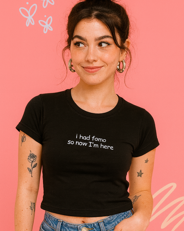A young woman with dark hair and tattoos on her arms is smiling and looking to the side against a pink background with white butterflies. She is wearing a black crop top with the text "I had somo so now I'm here" printed on it.