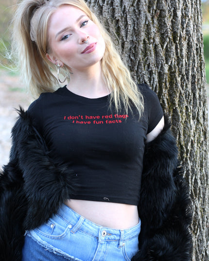 A young woman with blonde hair stands in front of a tree, wearing a black crop top with the text "I don't have red hair, I have fun facts" printed on it. She is also wearing a black fur coat and denim shorts.