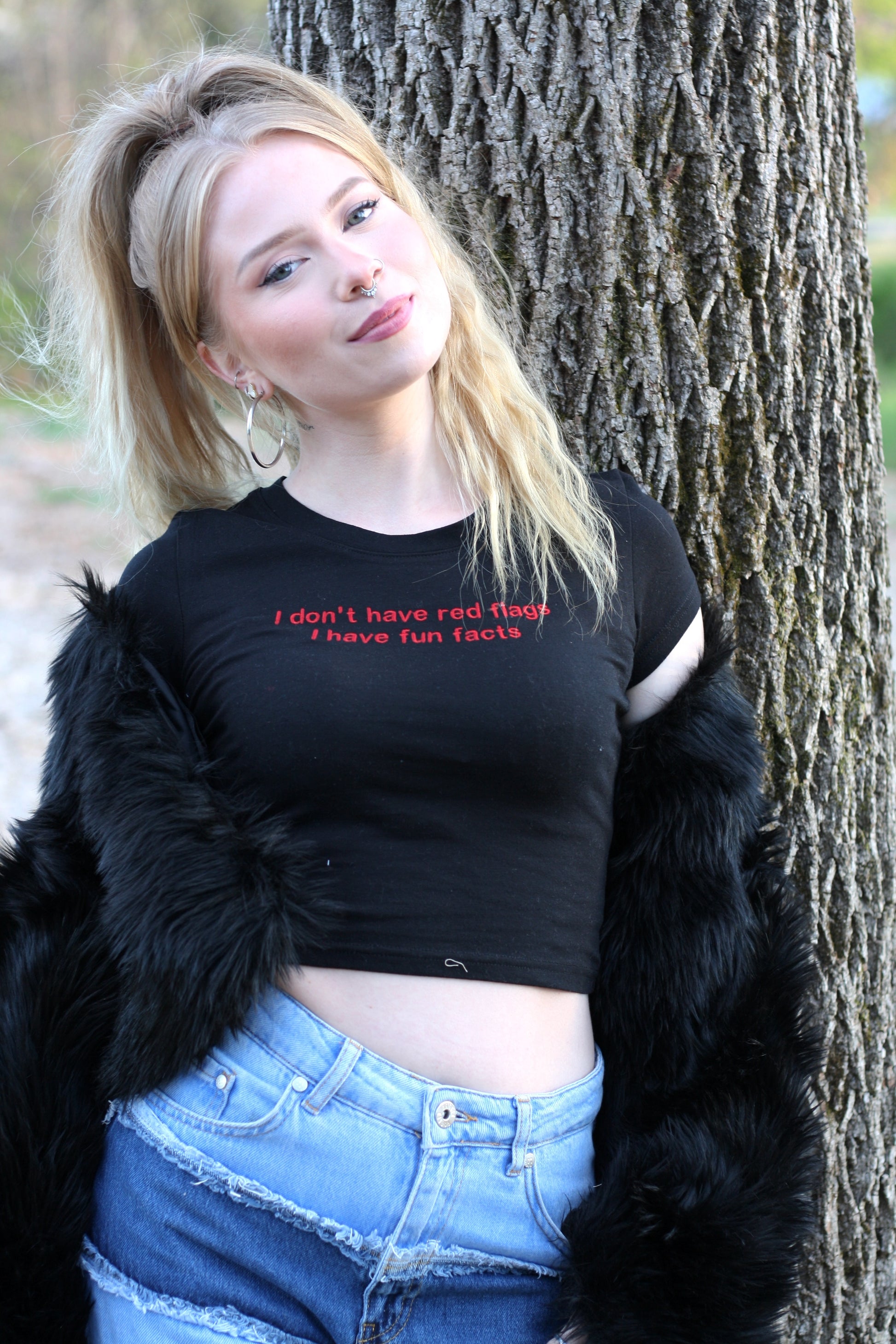 A young woman with blonde hair stands in front of a tree, wearing a black crop top with the text "I don't have red hair, I have fun facts" printed on it. She is also wearing a black fur coat and denim shorts.