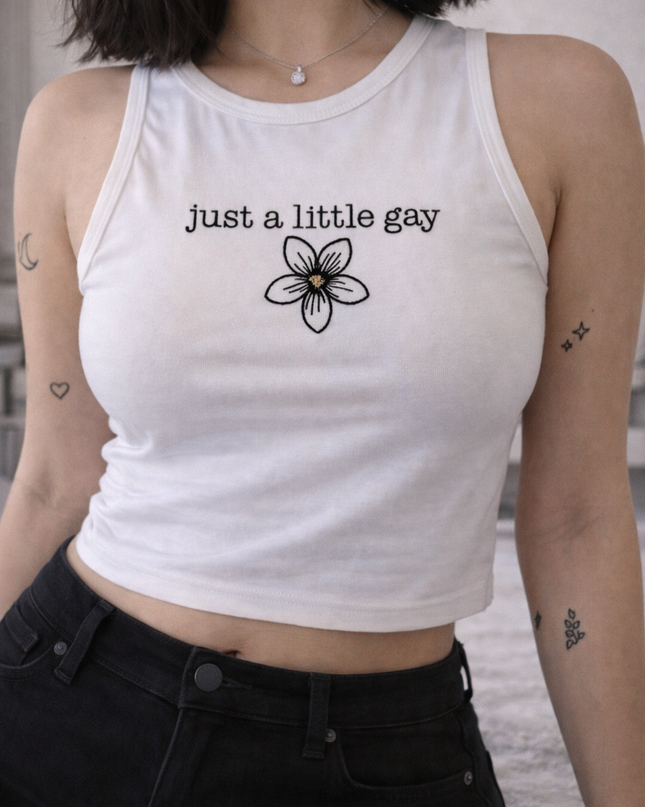 A woman wearing a white crop top with the text "Just a little gay" printed on it, standing in front of a plain background.