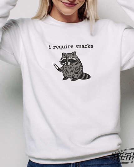 A woman wearing a white sweatshirt with a graphic of a raccoon and the text "I require snacks" printed on it.