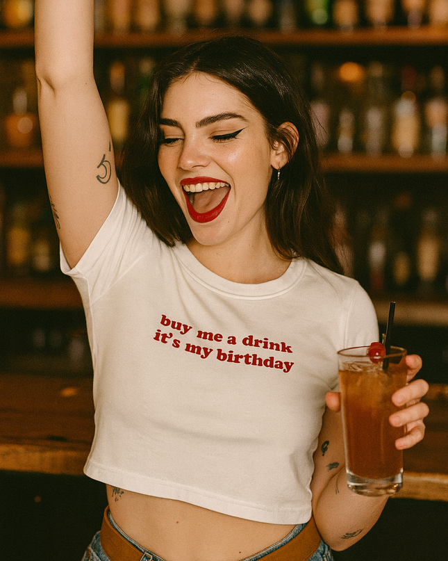 A woman wearing a white t-shirt with the text "buy me a drink it's my birthday" is holding a drink in her hand and raising her arm in a celebratory gesture.