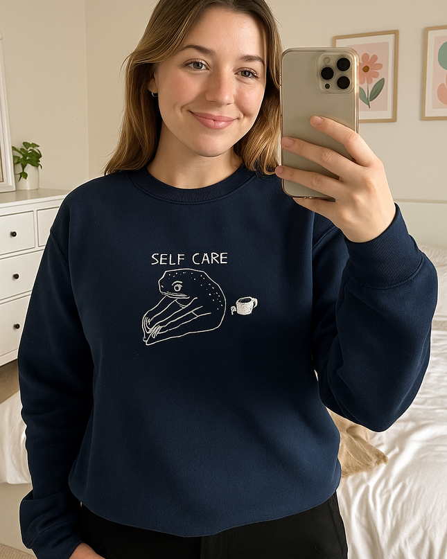 A woman is taking a selfie in a bedroom, wearing a navy blue sweatshirt with a white graphic and text that says "SELF CARE".