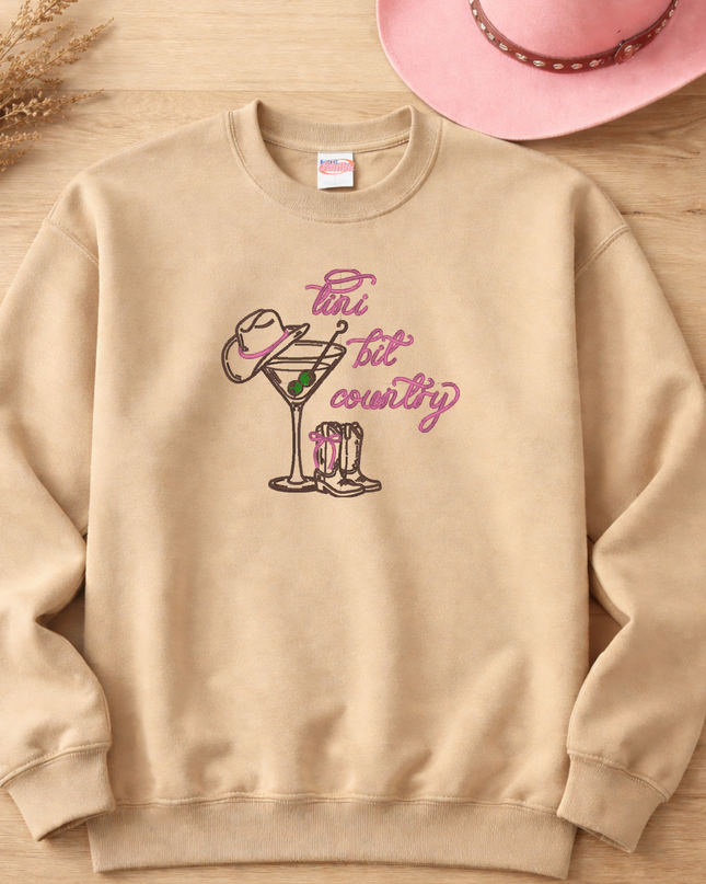 A beige sweatshirt with a graphic design of a cocktail glass and a cowboy hat printed on it, along with the text "Pin & Tip Country" printed in pink.