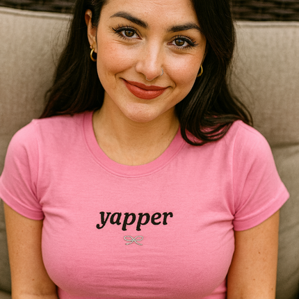 A young woman with long dark hair is sitting on a chair, wearing a pink t-shirt with the word "yapper" printed on it. She is smiling and looking directly at the camera.