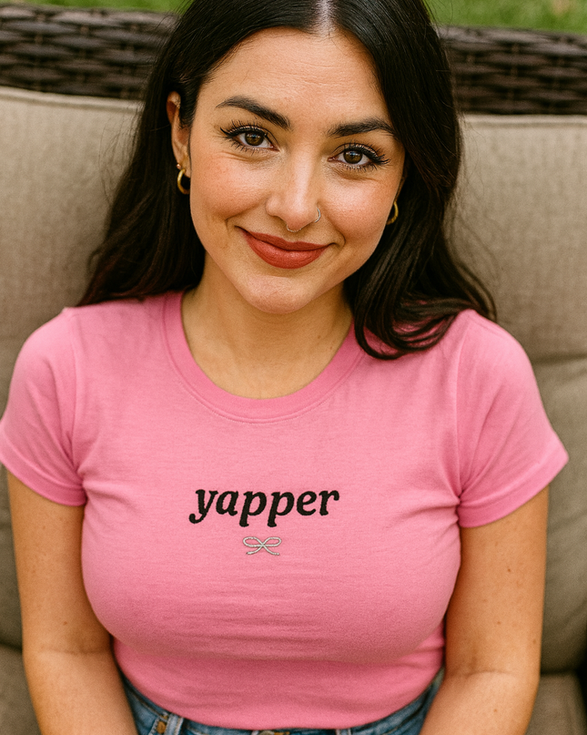 A young woman with long dark hair is sitting on a chair, wearing a pink t-shirt with the word "yapper" printed on it. She is smiling and looking directly at the camera.