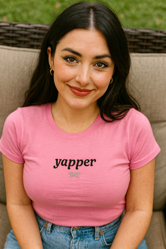 A young woman with long dark hair is sitting on a chair, wearing a pink t-shirt with the word "yapper" printed on it. She is smiling and looking directly at the camera.