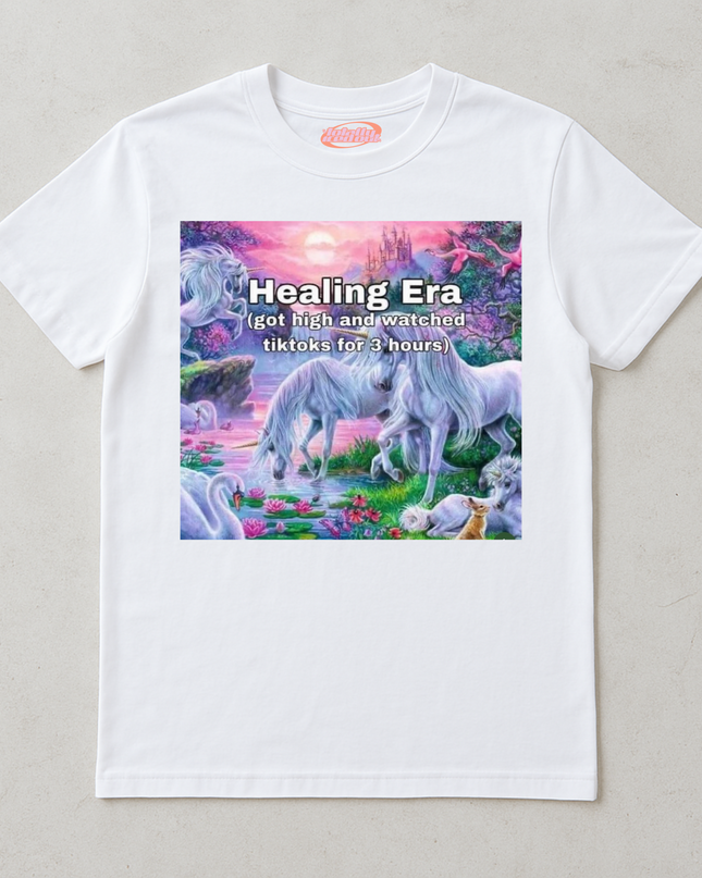A white t-shirt with a colorful graphic design depicting a unicorn surrounded by various fantastical elements, such as a castle, flowers, and a sunset.