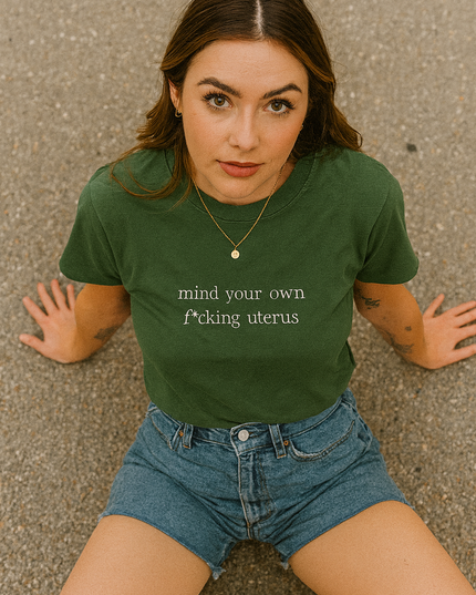 A young woman wearing a green t-shirt with the text "mind your own shit" and denim shorts is sitting on the ground.