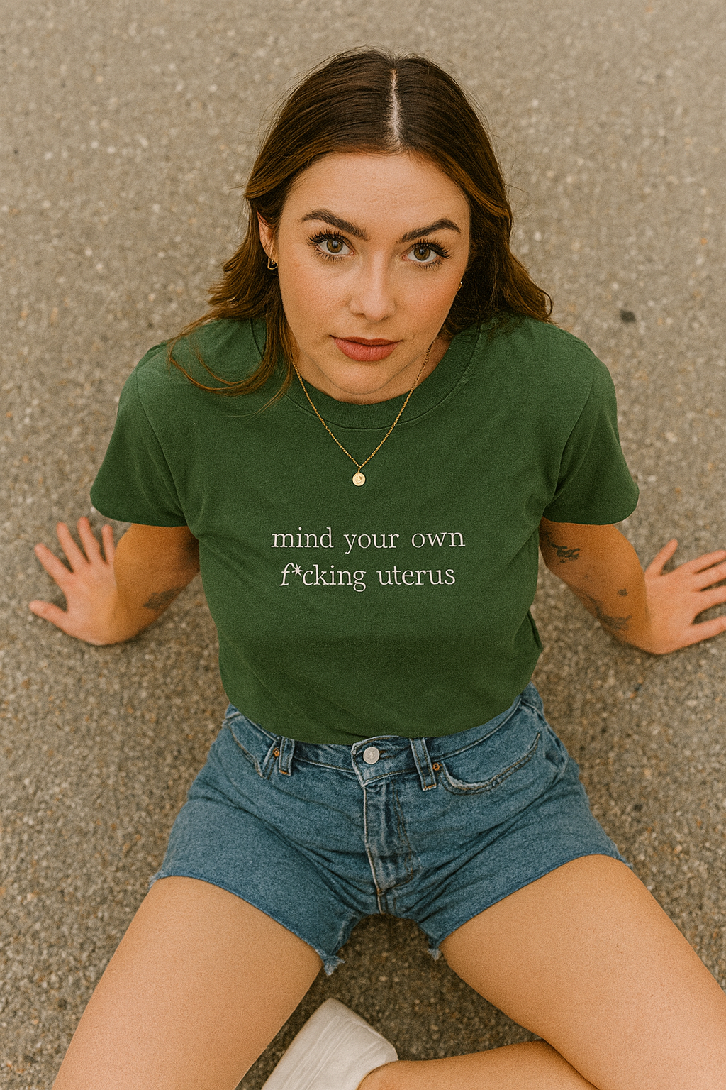 A young woman wearing a green t-shirt with the text "mind your own shit" and denim shorts is sitting on the ground.