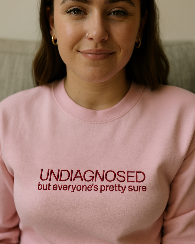 A woman wearing a pink sweatshirt with the text "UNDIAGNOSED but everyone's pretty sure" printed on it.