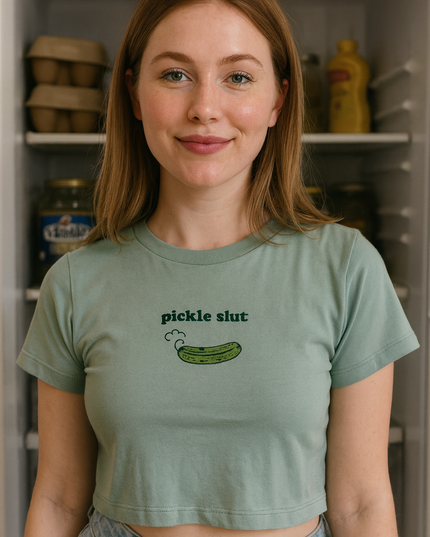A young woman with long brown hair is standing in front of an open refrigerator, wearing a green crop top with a graphic of a pickle on it.