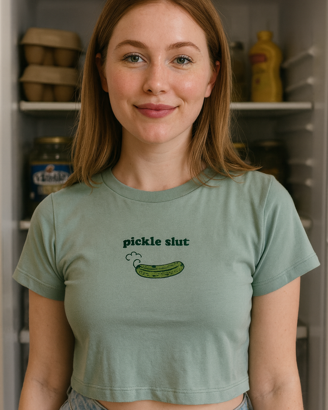 A young woman with long brown hair is standing in front of an open refrigerator, wearing a green crop top with a graphic of a pickle on it.