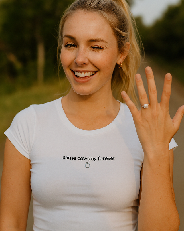 A smiling woman wearing a white t-shirt with the text "same cowboy forever" is standing outdoors, with trees and a road in the background.