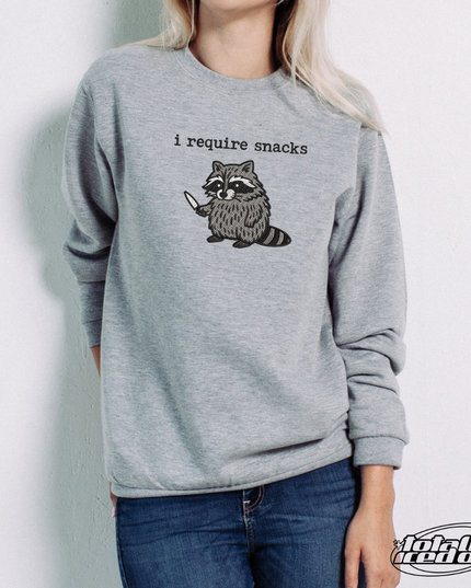 A person wearing a gray sweatshirt with a graphic of a raccoon holding a knife and the text "I require snacks" printed on it.