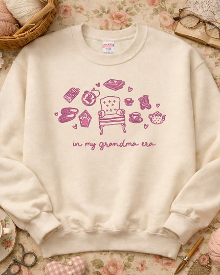 A beige sweatshirt with a graphic design of a chair surrounded by various objects, including a teapot, a cup, and a basket of yarn, along with the text "in my grandma era".