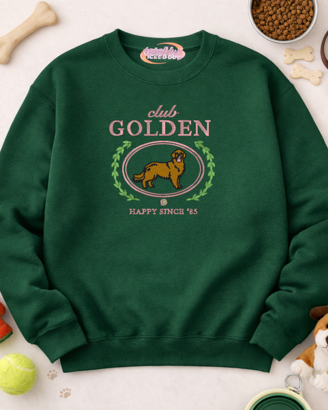 A green sweatshirt with a graphic of a golden retriever dog and the text "club GOLDEN" printed on it, along with the text "HAPPY SINCE '85". The sweatshirt is surrounded by various dog toys and accessories, including a bowl of dog food.
