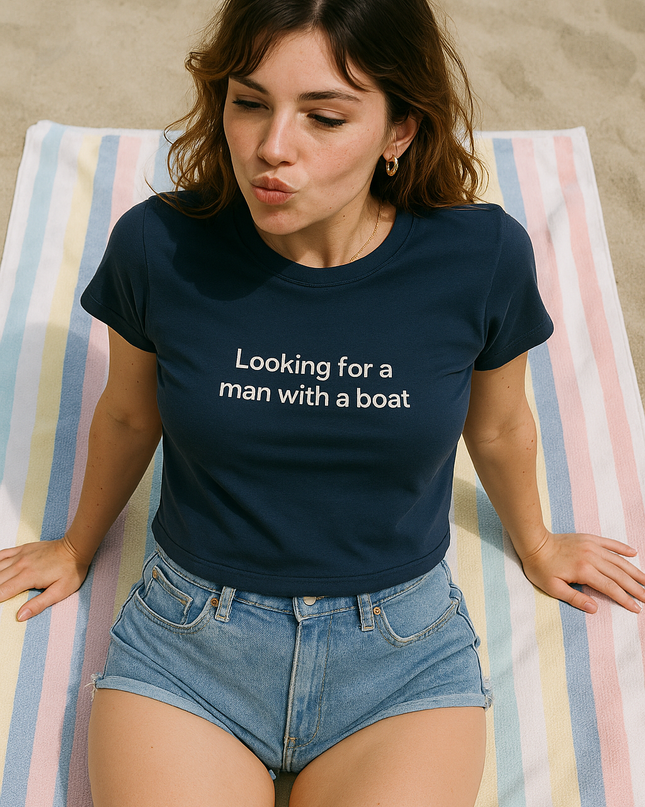 A woman sitting on a striped towel, wearing a blue t-shirt with the text "Looking for a man with a boat" and denim shorts.