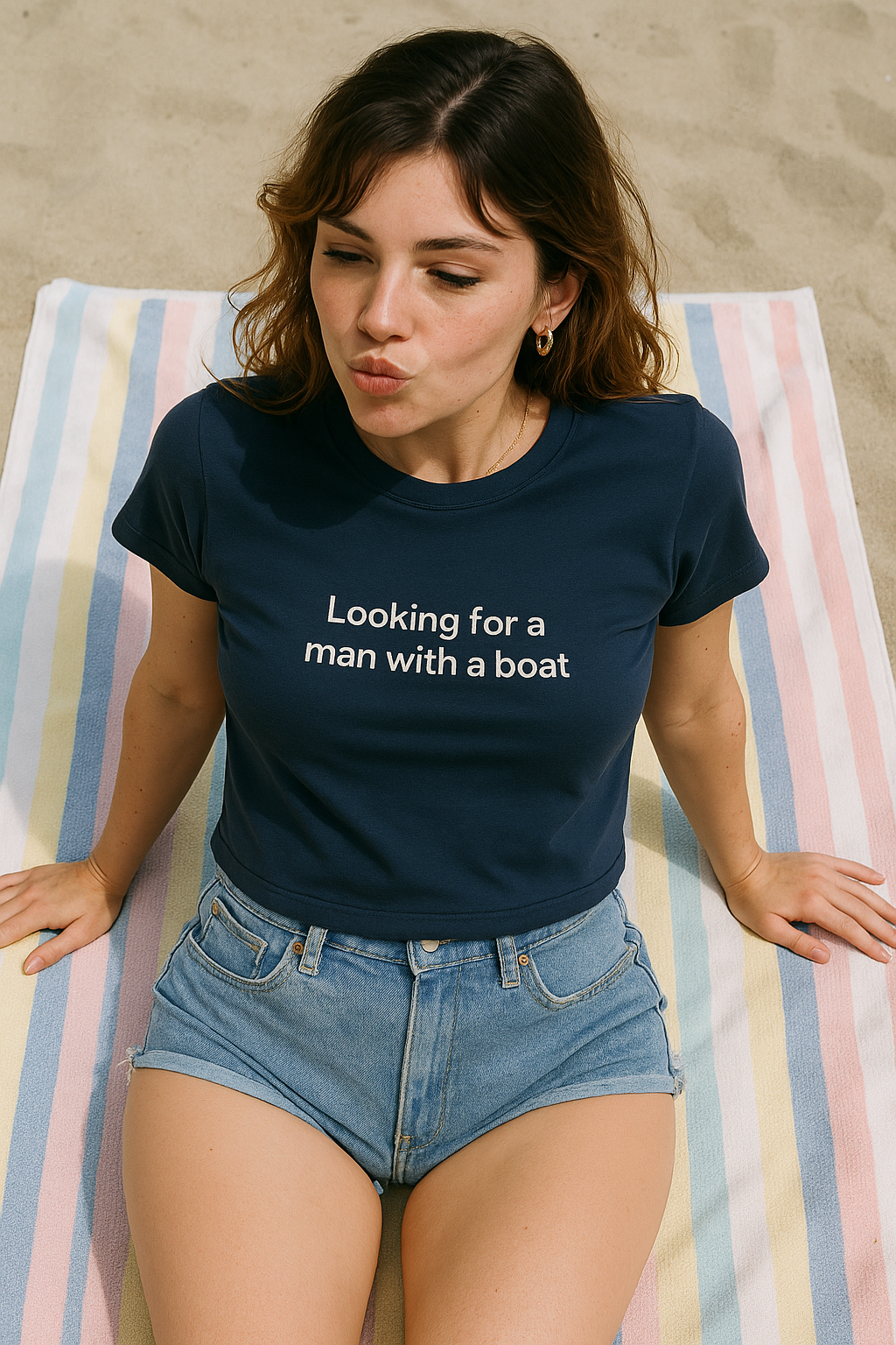 A woman sitting on a striped towel, wearing a blue t-shirt with the text "Looking for a man with a boat" and denim shorts.