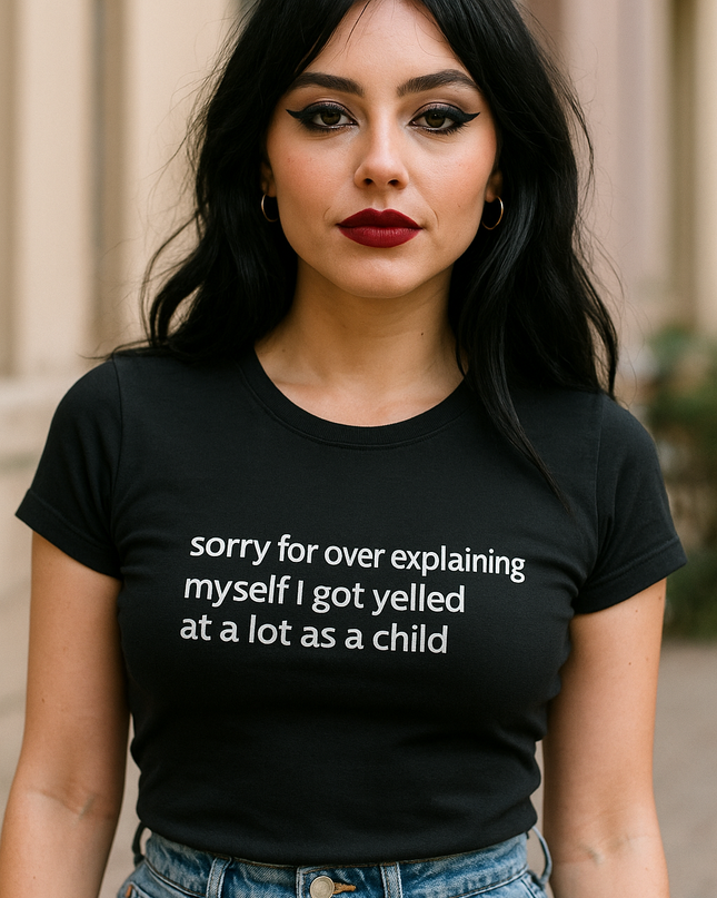 A woman with long dark hair wearing a black t-shirt with white text that says "Sorry for over explaining myself I got yelled at a lot as a child".