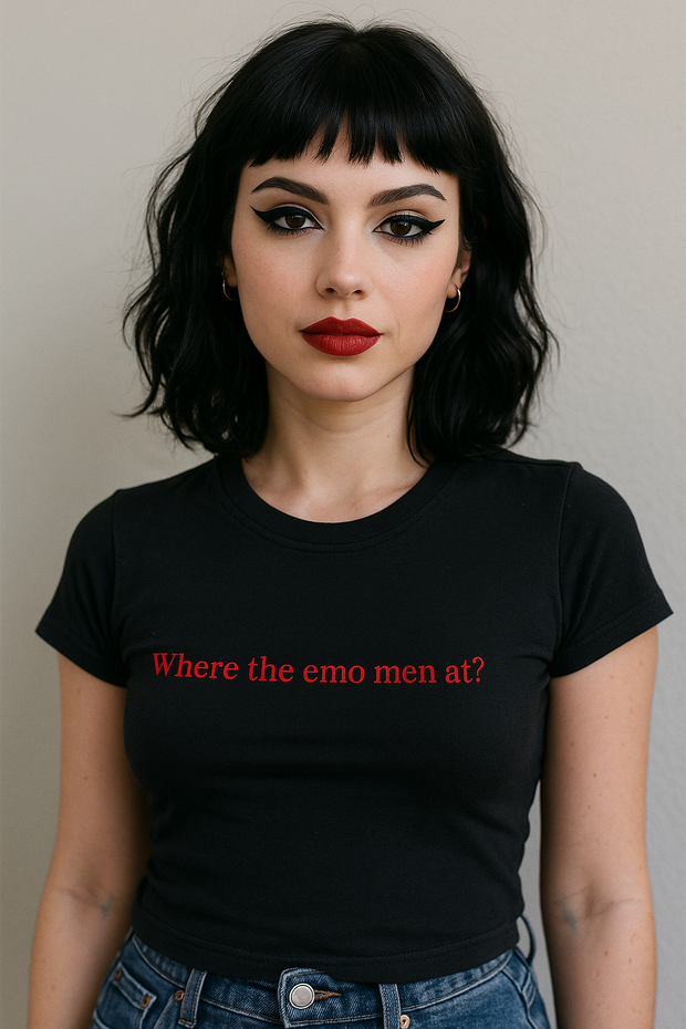 A woman with short, dark hair and bold red lipstick is wearing a black t-shirt with the text "Where the emo men at?" printed on it.