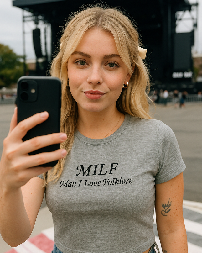 A young woman with blonde hair is taking a selfie with a black phone, wearing a gray t-shirt with the text "MILF Man I Love Folklore".