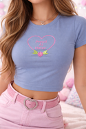 A woman wearing a blue crop top with a heart-shaped embroidered phrase and a pink belt buckle.