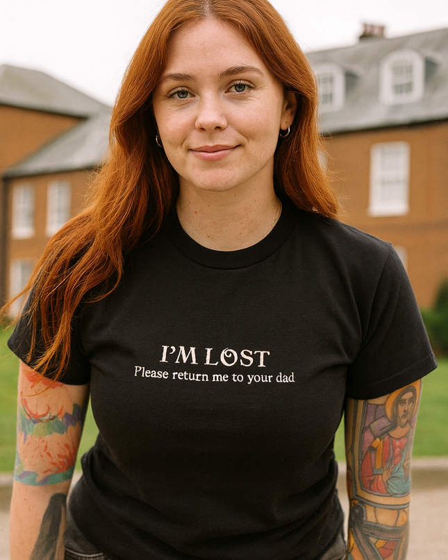 A young woman with long red hair wearing a black t-shirt with the text "I'm lost Please return me to your dad" stands in front of a brick building.