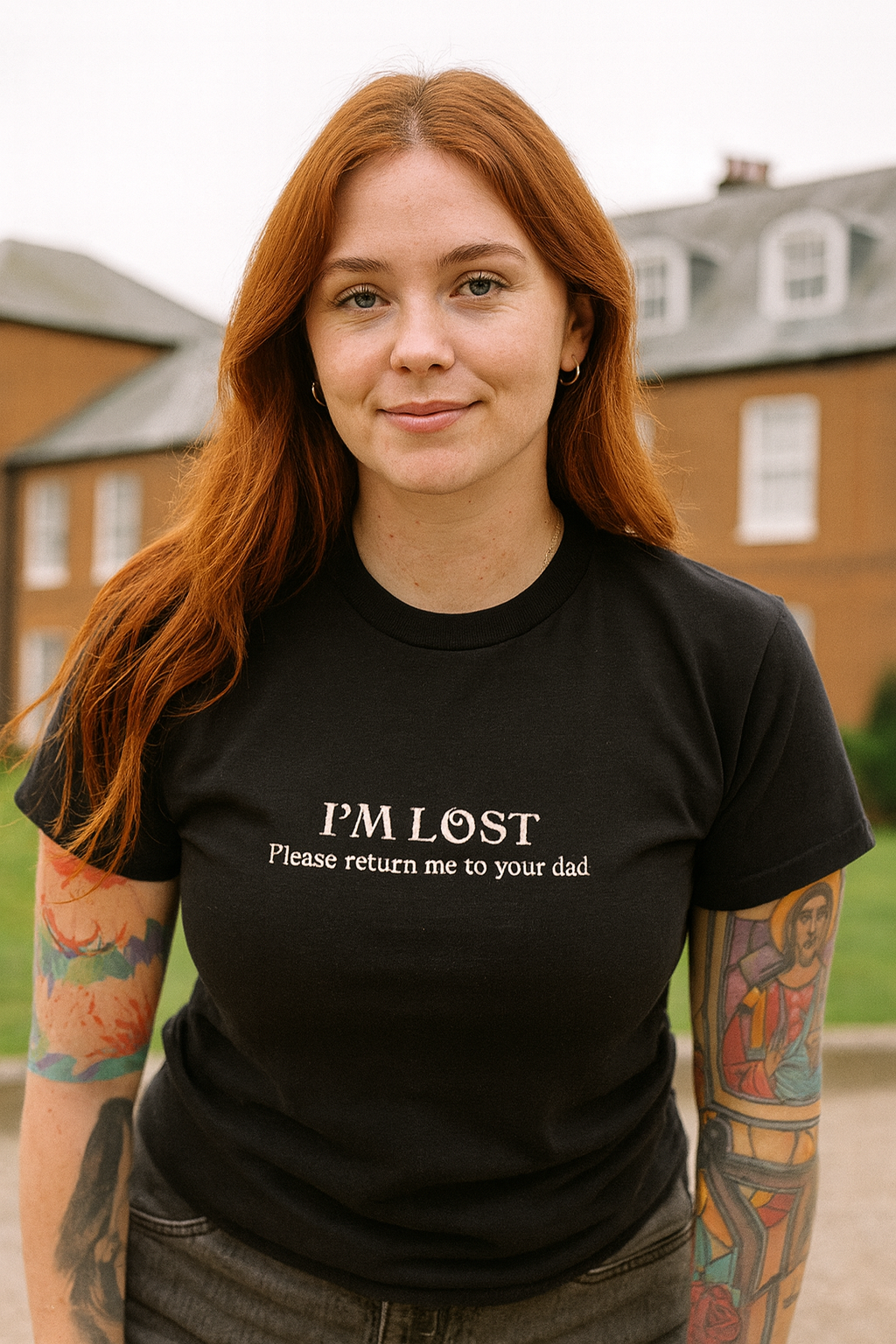 A young woman with long red hair wearing a black t-shirt with the text "I'm lost Please return me to your dad" stands in front of a brick building.