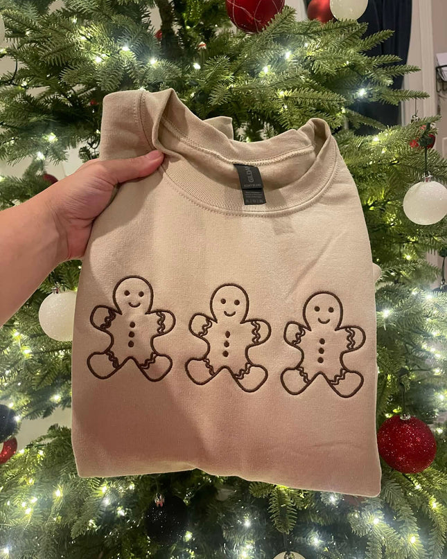 A hand is holding up a beige sweater with three gingerbread man designs on it, set against a backdrop of a Christmas tree decorated with ornaments.