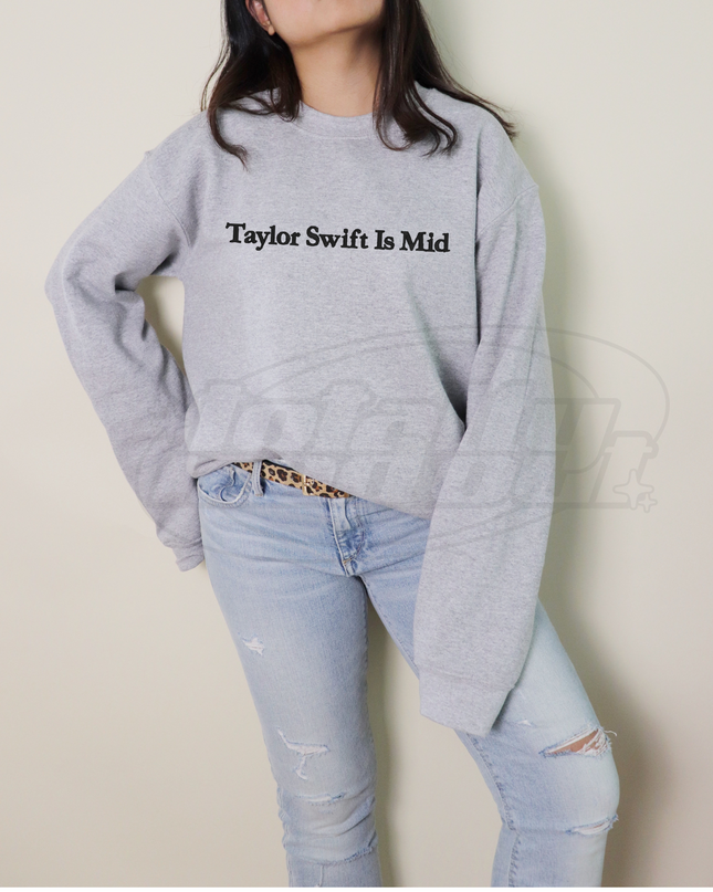 A person wearing a grey sweatshirt with the text "Taylor Swift Is Mid" printed on it, paired with distressed blue jeans.