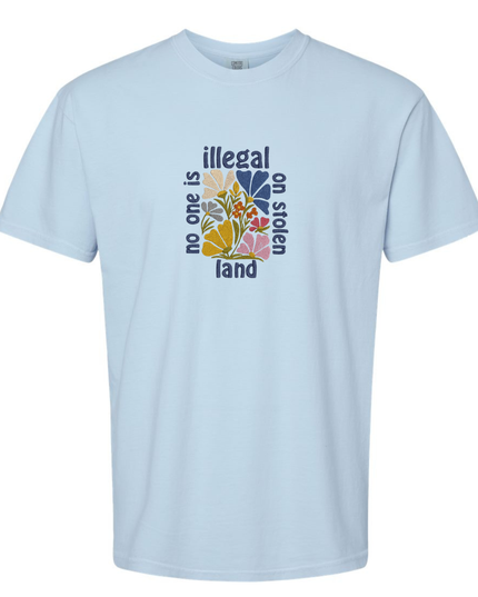A light blue t-shirt with a graphic design featuring a colorful flower-like shape surrounded by the text "Illegal on stolen land".
