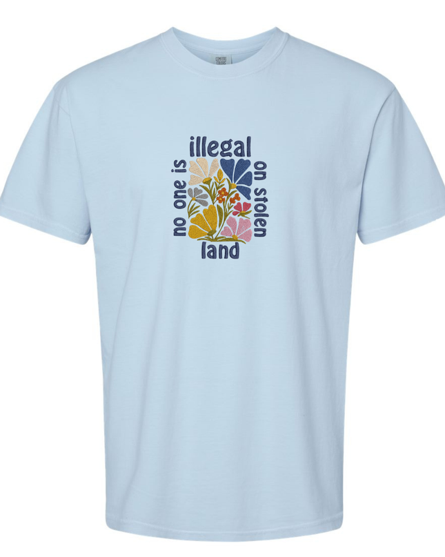A light blue t-shirt with a graphic design featuring a colorful flower-like shape surrounded by the text "Illegal on stolen land".