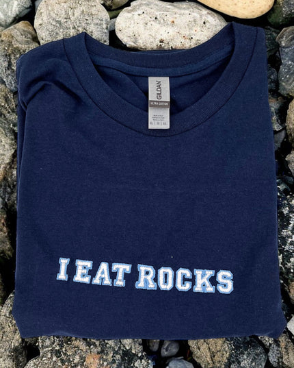 A navy blue t-shirt with the text "I EAT ROCKS" printed on it, laid out on a bed of rocks.