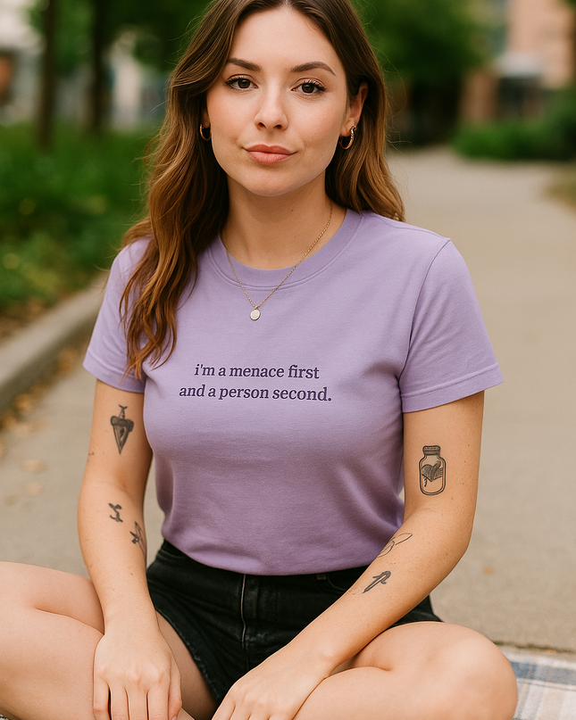 A young woman with long brown hair sits on the ground, wearing a purple t-shirt with text and tattoos on her arms.