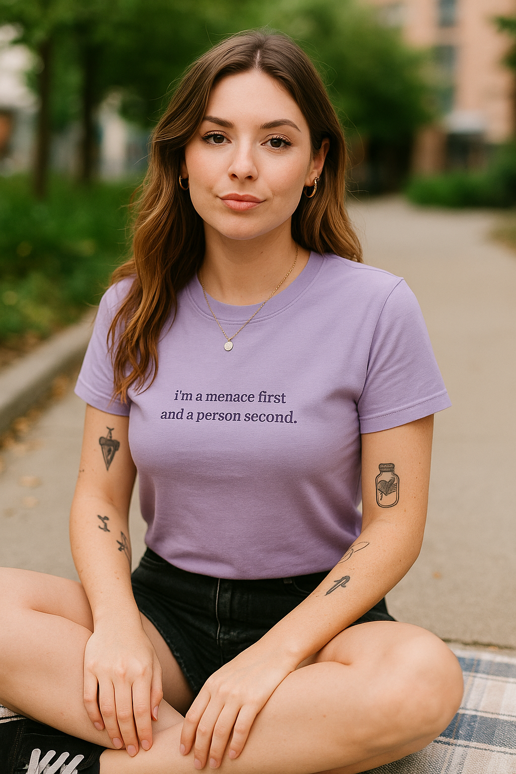 A young woman with long brown hair sits on the ground, wearing a purple t-shirt with text and tattoos on her arms.