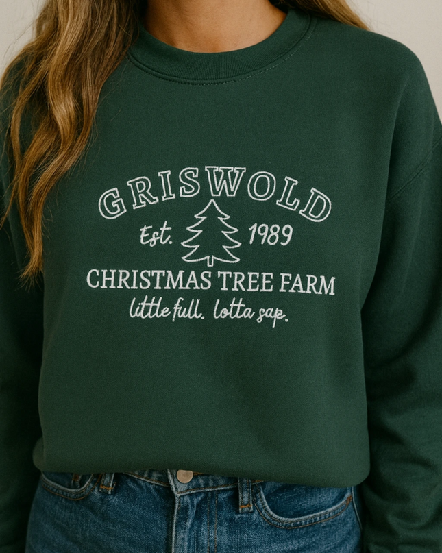 A woman wearing a green sweatshirt with white text that says "GRISWOLD est. 1989 CHRISTMAS TREE FARM little full, lotta lap."